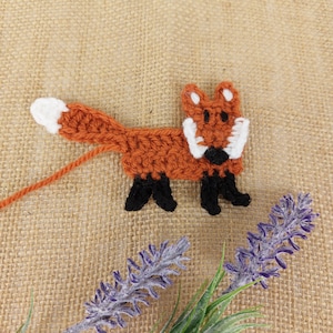 May include: A crocheted fox applique with orange fur, white accents, and black feet. The fox is on a hessian background.