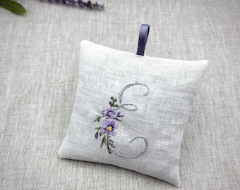 May include: A square light gray linen sachet featuring an embroidered letter C in a cursive font, accented with small purple floral details. A dark purple ribbon loop is attached to the top edge for hanging, and a sprig of dried lavender is nearby.