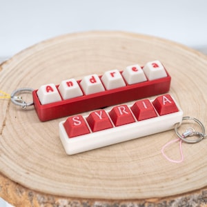 Peut inclure: Deux porte-clés avec les noms "Andrea" et "Syria", faits à partir de touches de clavier rouges et blanches. Le porte-clés "Andrea" est rouge et le porte-clés "Syria" est blanc. Les deux ont des porte-clés et sont sur une surface en bois.