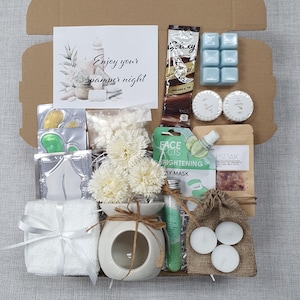 May include: A gift box filled with self-care items. Includes a white ceramic oil diffuser with flowers, a small white towel tied with a ribbon, candles, bath products, and a card that reads "Enjoy your pamper night."