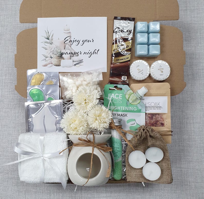 May include: A gift box filled with self-care items. Includes a white ceramic oil diffuser with flowers, a small white towel tied with a ribbon, candles, bath products, and a card that reads "Enjoy your pamper night."