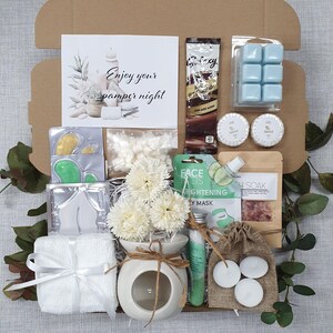 May include: A gift box filled with self-care items. Includes a white towel tied with a ribbon, a small ceramic oil diffuser with flowers, face masks, bath salts, candles, and a chocolate bar. The card reads "Enjoy your pamper night."