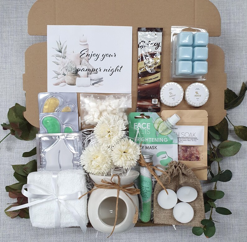 May include: A gift box filled with self-care items. Includes a white towel tied with a ribbon, a small ceramic oil diffuser with flowers, face masks, bath salts, candles, and a chocolate bar. The card reads "Enjoy your pamper night."