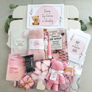 May include: A pink and white gift box filled with various items, including a pink bathrobe, a pink bath bomb, a pink face mask, a pink foot scrub, a pink candle, a pink teddy bear, and a set of baby milestone cards. The box also includes a card that says "Raising tiny humans is exhausting".
