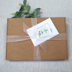 May include: A brown cardboard gift box tied with a sheer white ribbon and bow. A white card with the text "gift for You" is attached. Greenery is placed on top of the box.