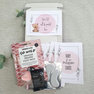 May include: A pink and white gift box with a pink bear illustration on the lid. The box contains a foot mask, a candle, eye masks, and baby milestone cards. The text "Your text will be printed here" is on the lid of the box. The text "BABY milestone CARDS" is on one of the cards.