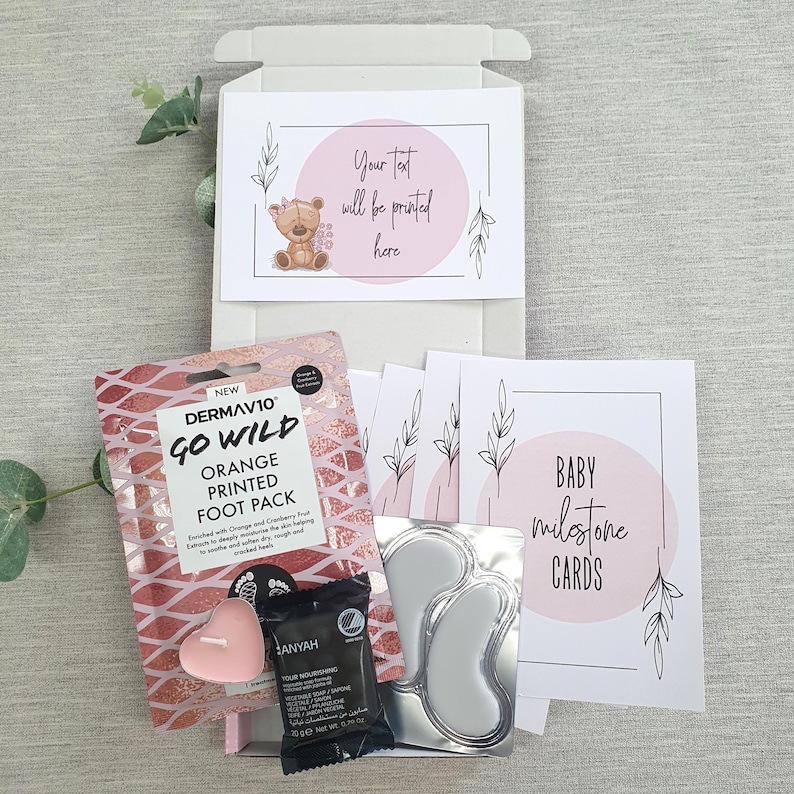 May include: A pink and white gift box with a pink bear illustration on the lid. The box contains a foot mask, a candle, eye masks, and baby milestone cards. The text "Your text will be printed here" is on the lid of the box. The text "BABY milestone CARDS" is on one of the cards.