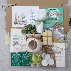 May include: A spa-themed gift box filled with self-care items. Includes a white ceramic oil diffuser, bath soak, face mask, candles, and a rolled white towel tied with a green ribbon. The box also contains tea bags and small bottles of shampoo and conditioner.