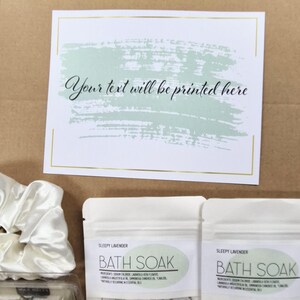 May include: A gift set with a white card featuring the text "Your text will be printed here" above a green brushstroke design. Two "Sleepy Lavender Bath Soak" packets and a white satin scrunchie are visible.