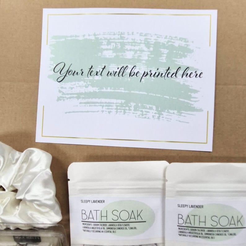 May include: A gift set with a white card featuring the text "Your text will be printed here" above a green brushstroke design. Two "Sleepy Lavender Bath Soak" packets and a white satin scrunchie are visible.