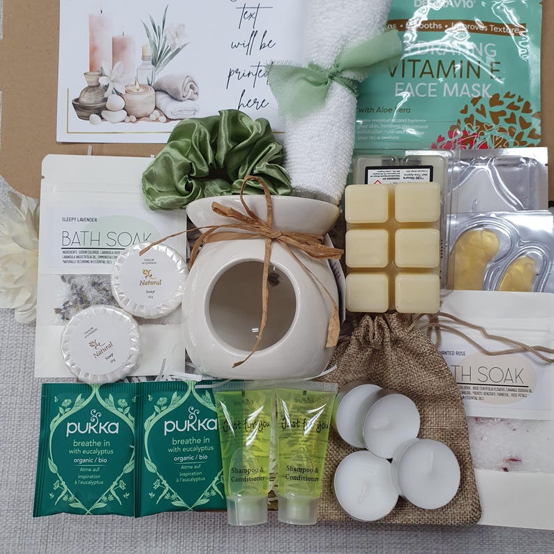 May include: A spa-themed gift set featuring a white ceramic oil diffuser tied with twine, a green scrunchie, a white towel with a green ribbon, and various bath and beauty products. Includes bath soak, soap, face mask, wax melts, tea lights, and shampoo and conditioner.