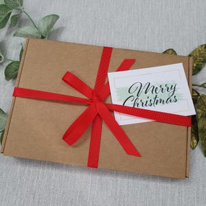 May include: A brown cardboard gift box tied with a bright red ribbon. A white card with the words "Merry Christmas" in elegant script is attached to the box. The box is decorated with greenery.