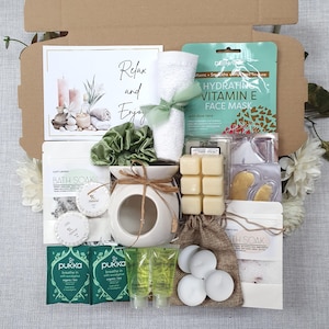 May include: A gift box filled with self-care items. Includes a face mask, bath soak, candles, a white towel tied with a green ribbon, a white ceramic oil diffuser, and a card that says "Relax and Enjoy".