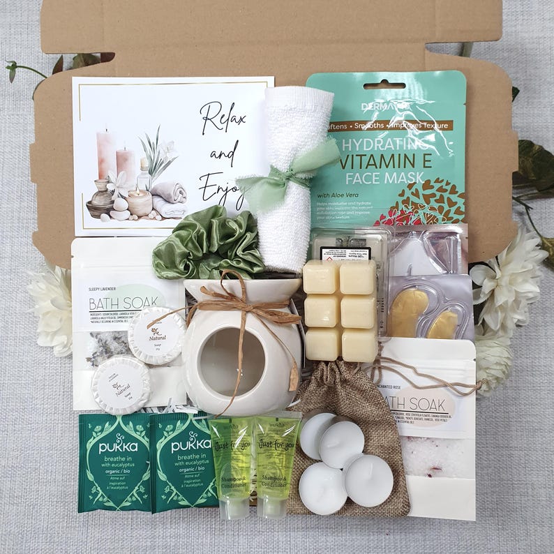 May include: A gift box filled with self-care items. Includes a face mask, bath soak, candles, a white towel tied with a green ribbon, a white ceramic oil diffuser, and a card that says "Relax and Enjoy".