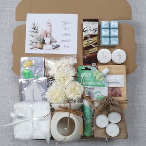 May include: A gift box filled with self-care items. Includes a card with text, a chocolate bar, blue wax melts, bath products, a face mask, a small diffuser with flowers, a towel, and tea light candles.