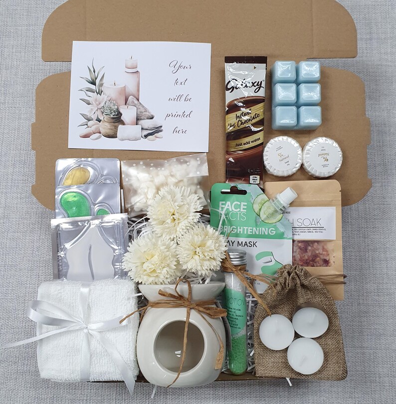 May include: A gift box filled with self-care items. Includes a card with text, a chocolate bar, blue wax melts, bath products, a face mask, a small diffuser with flowers, a towel, and tea light candles.