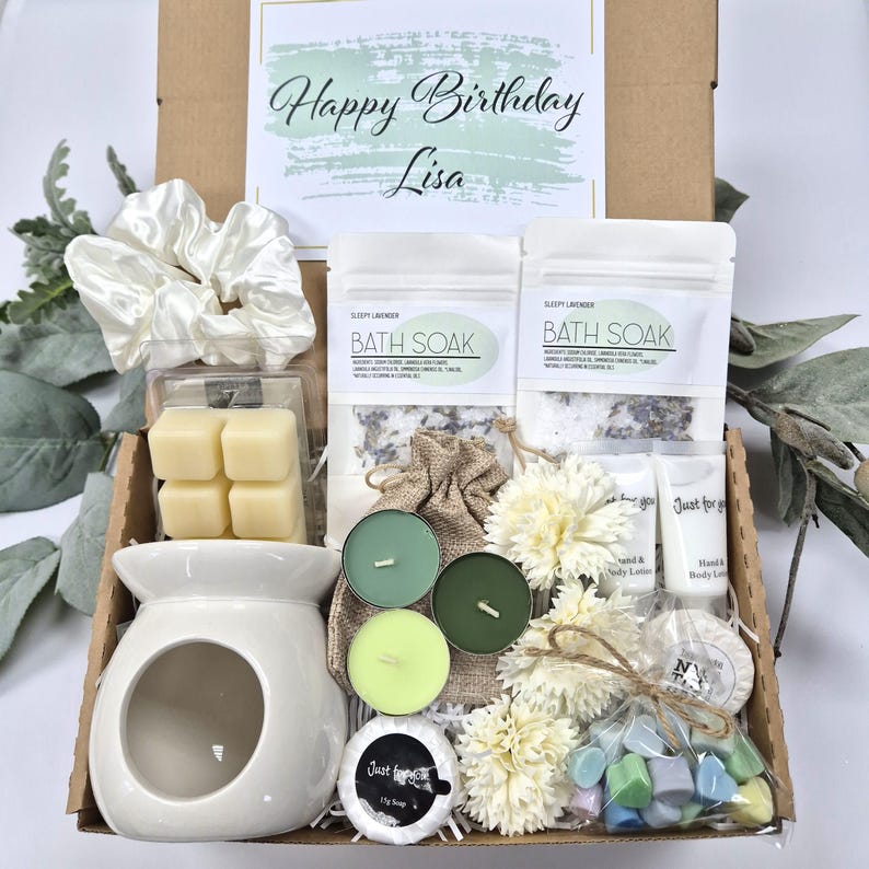 May include: A gift box filled with self-care items. Includes a white ceramic oil warmer, wax melts, bath soak, candles, soap, hand and body lotion, a scrunchie, and a decorative floral arrangement. The box has a card that says "Happy Birthday Lisa".