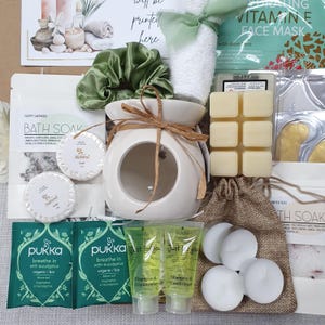 May include: A gift set with spa and relaxation items. Includes a white ceramic oil warmer tied with twine, a green satin scrunchie, a white towel, bath soak, soap, tea, shampoo, conditioner, candles, and a face mask.