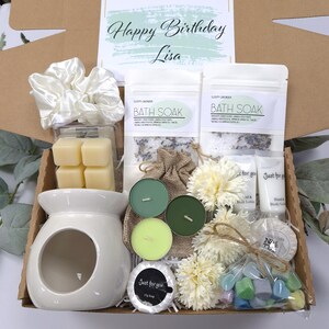 May include: A gift box with a variety of self-care items. It includes a white ceramic oil warmer, bath soak packets, candles, soap, and a scrunchie. The box also contains a card that says "Happy Birthday Lisa."