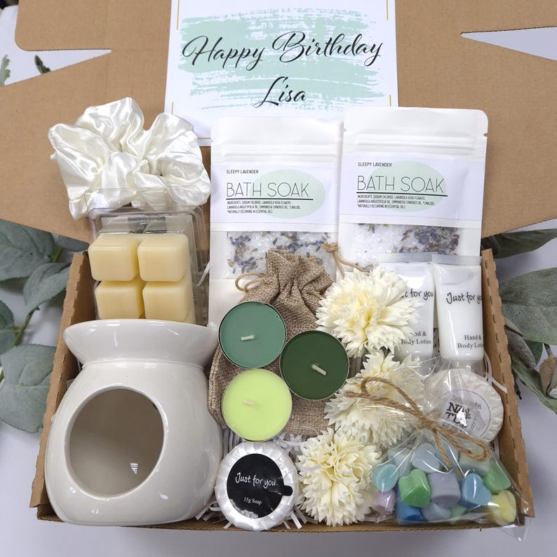 May include: A gift box with a variety of self-care items. It includes a white ceramic oil warmer, bath soak packets, candles, soap, and a scrunchie. The box also contains a card that says "Happy Birthday Lisa."