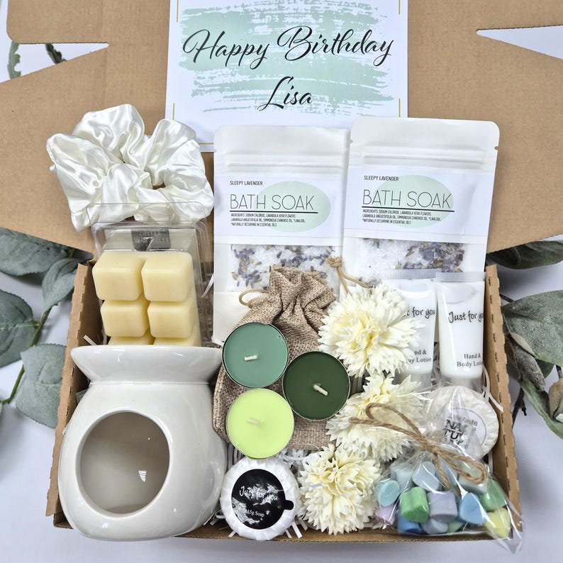 May include: A gift box filled with self-care items. Includes a white ceramic oil warmer, wax melts, bath soak, candles, lotion, and a scrunchie. The box is decorated with white flowers and a card that says "Happy Birthday Lisa."