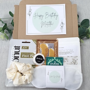 Może przedstawiać: Pudełko prezentowe na urodziny z kartką z napisem "Happy Birthday Matilda!" i r&oacute;żnymi produktami do pielęgnacji. Pudełko zawiera maseczkę do st&oacute;p, gumkę do włos&oacute;w, satynową maskę na oczy, s&oacute;l do kąpieli i inne przedmioty.