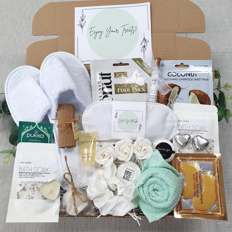 Może przedstawiać: Zestaw prezentowy do spa wypełniony artykułami do pielęgnacji. Zawiera białe kapcie, maskę do spania, produkty do kąpieli, maskę na twarz, zieloną ściereczkę i kartę z napisem "Enjoy Your Treats!"