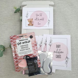 May include: A pink and white gift box with a teddy bear illustration and the text "Your text will be printed here". The box contains a foot mask, a candle, a small jar of cream, and a set of baby milestone cards with pink and white floral designs.