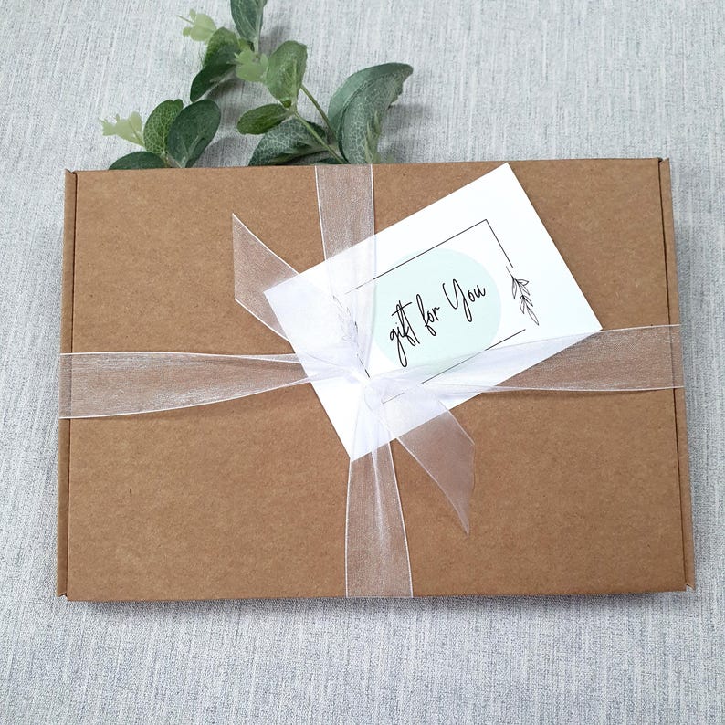 May include: A brown cardboard gift box tied with a sheer white ribbon. A white card with the text "gift for You" is attached to the ribbon. Greenery is placed on top of the box.