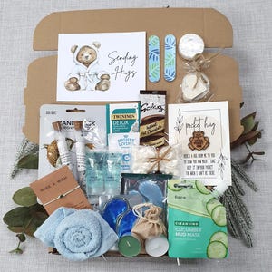 Może przedstawiać: Pudełko prezentowe zawierające kartkę Sending Hugs, dwa pilniki do paznokci, przedmiot w kształcie serca, maskę do rąk, herbatę Twinings, gorącą czekoladę Galaxy, kartkę pocket hug z drewnianym misiem, sole do kąpieli, mini balsamy, pianki marshmallow, niebieskie maski pod oczy, bransoletkę, niebieski ręcznik, świece i maskę błotną z ogórkiem.