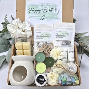 May include: A gift box with a variety of self-care items. Includes a white ceramic oil warmer, candles, bath soak, soap, hand and body lotion, wax melts, a scrunchie, and a card that says "Happy Birthday Lisa."
