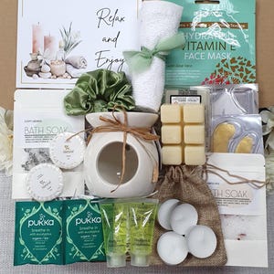 May include: A gift box filled with self-care items. Includes a white ceramic oil diffuser, bath soak, face mask, bath bombs, and Pukka tea. The box also contains a green scrunchie and a rolled white towel tied with a green ribbon. The text "Relax and Enjoy" is visible.