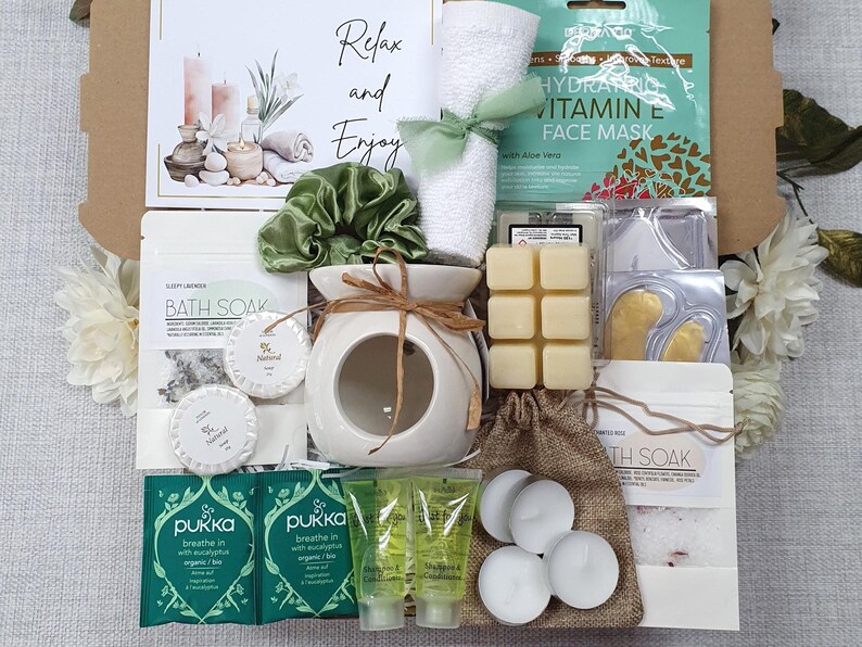 May include: A gift box filled with self-care items. Includes a white ceramic oil diffuser, bath soak, face mask, bath bombs, and Pukka tea. The box also contains a green scrunchie and a rolled white towel tied with a green ribbon. The text "Relax and Enjoy" is visible.
