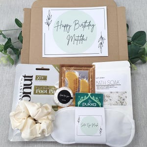 Może przedstawiać: Pudełko prezentowe na urodziny z kartką z napisem "Happy Birthday Matilda!" i r&oacute;żnymi produktami do pielęgnacji. Pudełko zawiera maseczkę do st&oacute;p, gumkę do włos&oacute;w, satynową maskę na oczy, s&oacute;l do kąpieli i inne przedmioty.