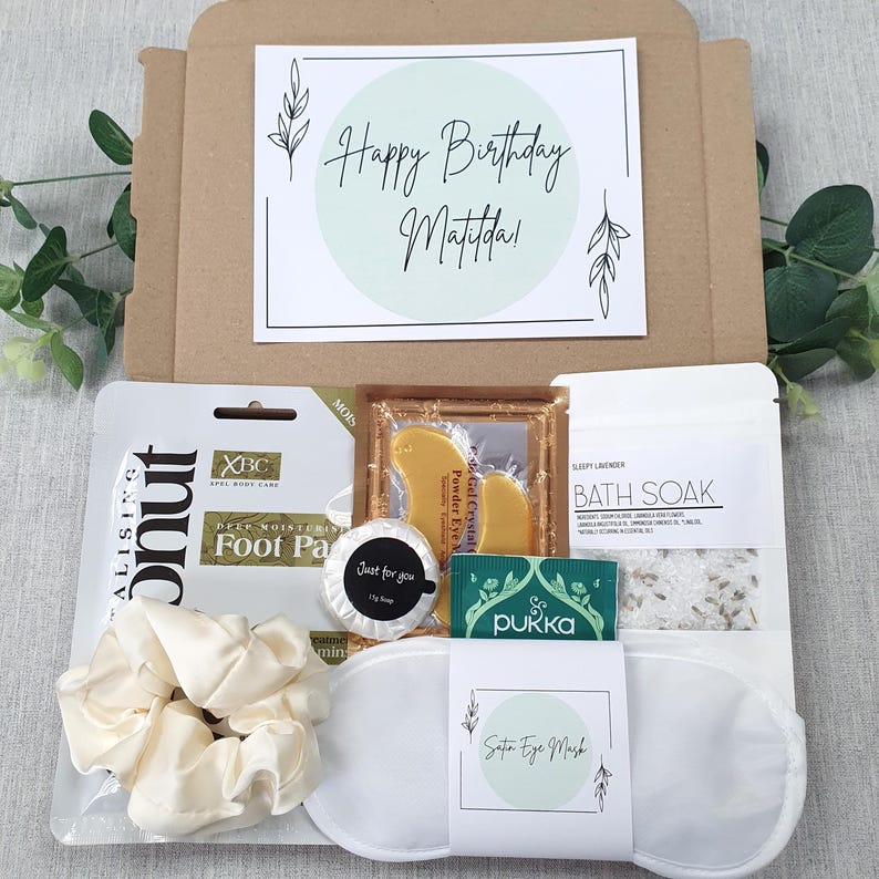 Może przedstawiać: Pudełko prezentowe na urodziny z kartką z napisem "Happy Birthday Matilda!" i r&oacute;żnymi produktami do pielęgnacji. Pudełko zawiera maseczkę do st&oacute;p, gumkę do włos&oacute;w, satynową maskę na oczy, s&oacute;l do kąpieli i inne przedmioty.