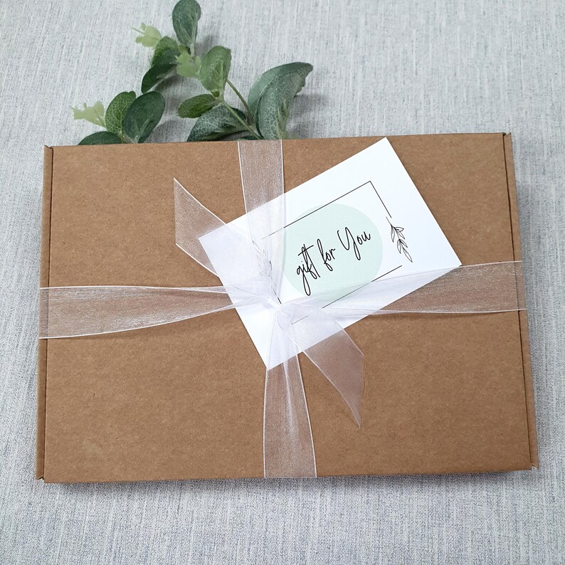 May include: A brown cardboard gift box tied with a sheer white ribbon. A white card with the words "gift for You" is attached to the ribbon. Greenery is placed on top of the box, creating a simple, elegant presentation.