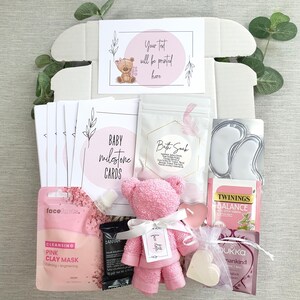 May include: A pink and white gift box filled with baby items. The box includes a pink teddy bear, bath soak, milestone cards, a face mask, tea, and a heart-shaped soap.