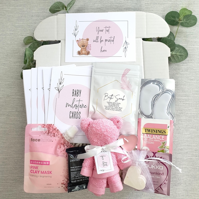 May include: A pink and white gift box filled with baby items. The box includes a pink teddy bear, bath soak, milestone cards, a face mask, tea, and a heart-shaped soap.