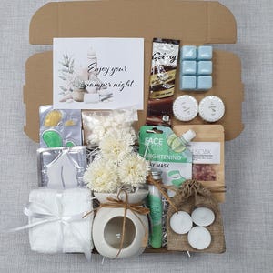May include: A gift box filled with self-care items. Includes a white towel tied with a ribbon, a ceramic oil diffuser with white flowers, and a card that reads "Enjoy your pamper night." Other items include face masks, bath salts, and candles.