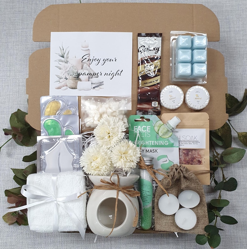 May include: A gift box filled with pampering items. Includes a card with text "Enjoy your pamper night", a chocolate bar, blue wax melts, soaps, eye masks, a diffuser with flowers, a face mask, bath soak, tea lights, and a small towel.