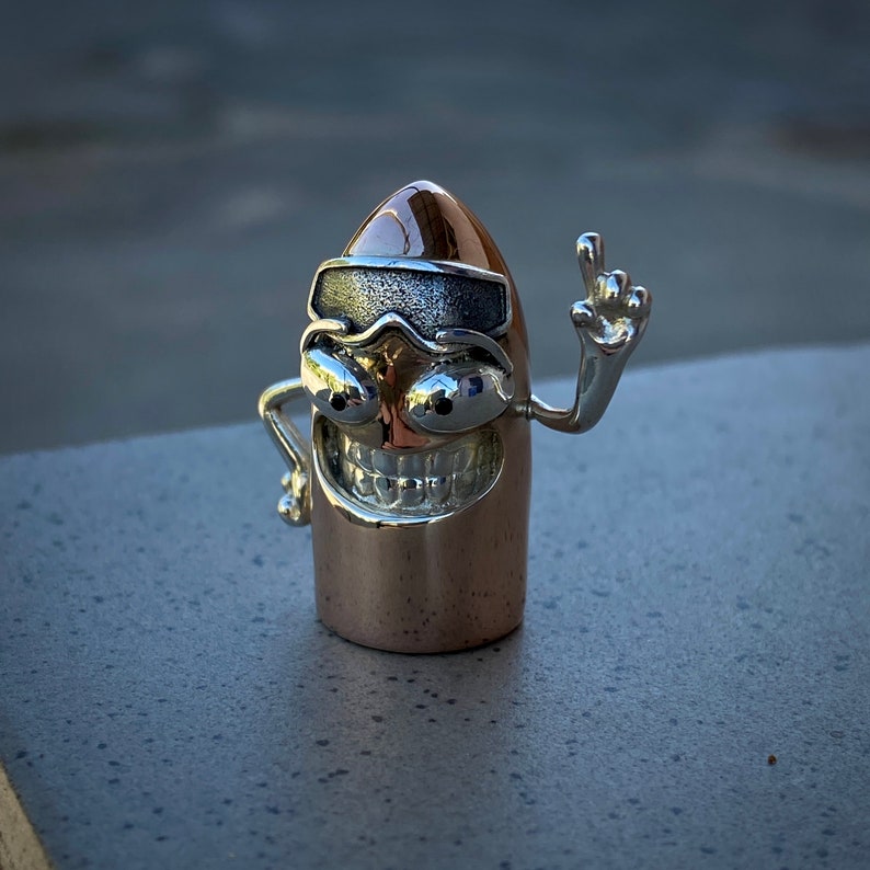 May include: A silver and copper-colored figurine of a bullet with a smiling face, wearing a silver eye patch and sunglasses, and raising one arm in the air.