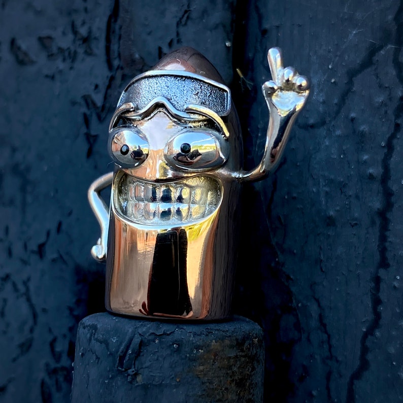May include: A silver and gold-toned figurine of a smiling character with a single raised arm and a pair of goggles. The figurine is shaped like a bullet and has a textured surface.