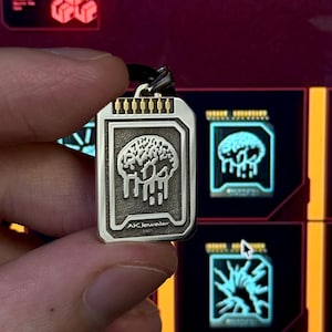 Könnte beinhalten: Ein rechteckiger silberner Anhänger in Form eines Datenchips mit einer erhabenen Gehirngrafik und tropfenden Details. Die Oberkante hat goldfarbene Kontaktstifte und die Unterseite ist mit dem Text AK Jeweler und der Zahl 925 graviert.