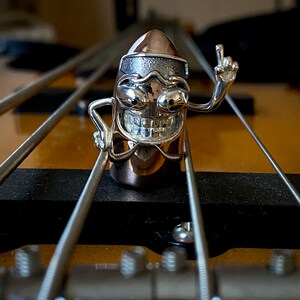 May include: A silver and gold bullet-shaped ring with a cartoon face and a raised middle finger. The ring is on a guitar neck.