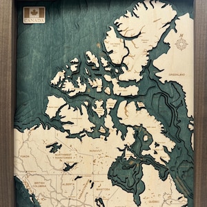 May include: A framed wooden map of Canada, featuring a layered design with teal waters and beige landmasses. The map includes the provinces and territories, with the Canadian flag in the upper left corner. The frame is a dark brown color.