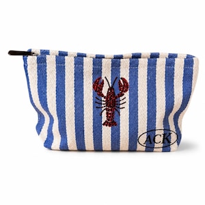 May include: A zippered pouch featuring a blue and white vertical striped pattern. The center displays a red lobster motif embellished with small crystals. The lower right corner features a black oval containing the text ACK in a serif font.
