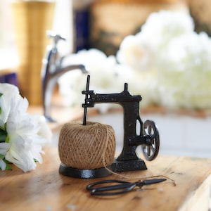 May include: A black metal twine dispenser shaped like a sewing machine with a spool of brown twine and a pair of black scissors on a wooden table.