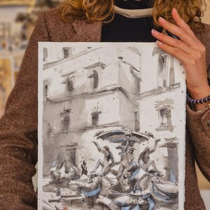 May include: Watercolor painting of a fountain with figures, in shades of gray and blue. The artwork depicts a detailed architectural scene with a water feature. The painting is held by a person.