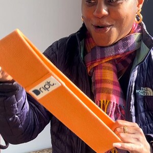 May include: A woman holds up an orange exercise band with the word "Dript" printed on it. The band is made of fabric and has a textured surface.