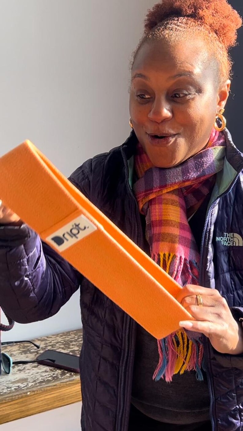May include: A woman holds up an orange exercise band with the word "Dript" printed on it. The band is made of fabric and has a textured surface.